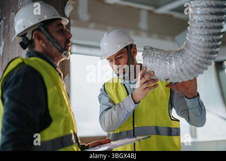 Two construction workers inspecting HVAC duct work in a commercial building wearing safety vests and helmets Stock Photo