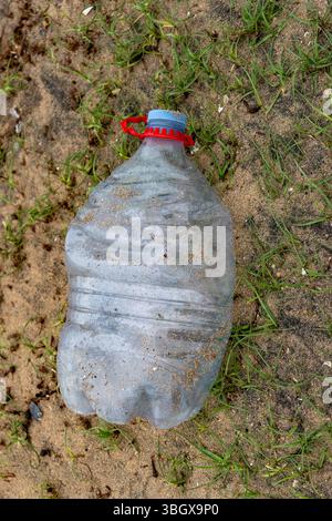 Plastic bottle is laying on the ground in a field. The bottle is empty and has a red cap. The bottle is surrounded by grass and dirt, giving it a some Stock Photo