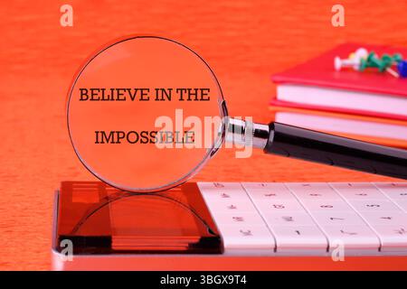 Motivational quote. BELIEVE IN THE IMPOSSIBLE text focused through a magnifying glass on an orange background, based on a calculator. Stock Photo