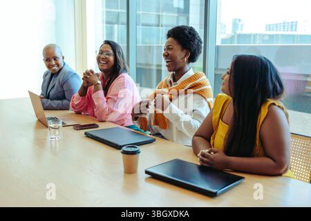 A diverse group of colleagues from a creative agency enjoying a lively discussion during a business meeting in a modern office with large windows, lap Stock Photo