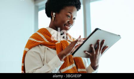 A woman is smiling as she uses a digital tablet in a modern office setting. She wears a bright, orange-striped sweater and wireless earbuds, creating Stock Photo