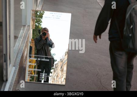 A photographer captures a creative self-portrait reflected in a mirror placed on an urban street, with a camera visible and a passerby blurred Stock Photo