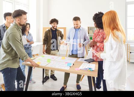 Multiracial group of young people standing around desk, working together in collaborative project. Stock Photo