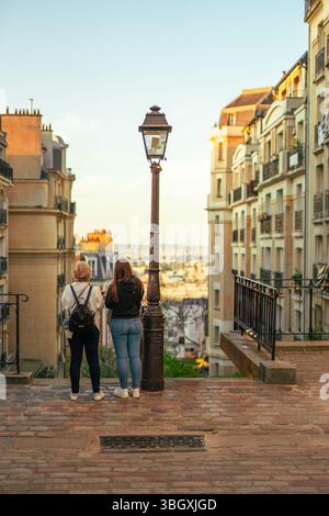 Seen from behind, two women stand together by a classic Parisian lamppost, admiring the expansive city view from the top of a hill in Montmartre durin Stock Photo