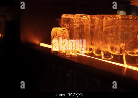 In a dark, atmospheric bar, a row of clean glasses sits on a shelf, glowing from the warm, orange light of an LED strip below. Stock Photo
