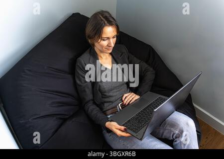 Relaxed female freelancer working on laptop from bean bag chair, top down view Stock Photo