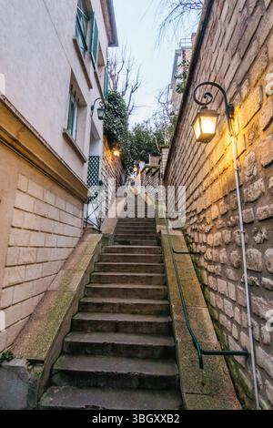 An atmospheric view looking up a narrow stone staircase squeezed between old buildings. The steps are illuminated by the warm glow of traditional lamp Stock Photo