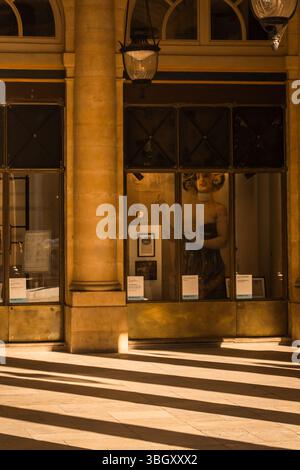 A shop window display featuring a mannequin, seen through an arched opening in a classic European arcade. Strong shadows from pillars stripe the floor Stock Photo
