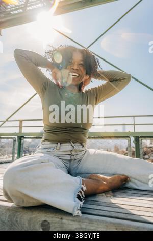 Smiling woman sits cross-legged in sunlight on NYC rooftop deck Stock Photo