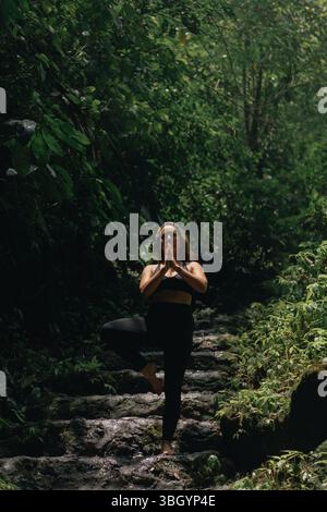 Woman practicing yoga pose in forest surrounded by lush greenery Stock Photo
