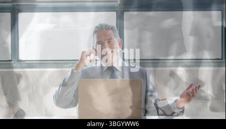 Smiling man in business suit holding smartphone to ear and gesturing at office desk, with laptop Stock Photo