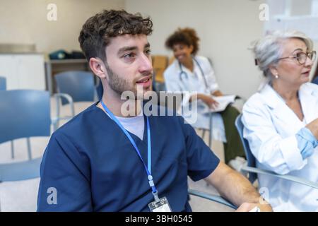 Focused young male nurse listening attentively during a medical conference, surrounded by colleagues in a hospital setting Stock Photo