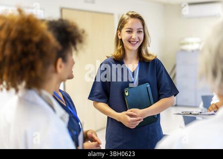 Happy nurse holding a tablet and smiling while engaging in a briefing with the medical team in a bustling hospital corridor Stock Photo