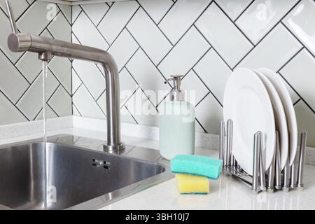 Running water from kitchen sink beside soap dispenser, stacked white plates in dish rack and sponges on tiled counter, dishwashing, daily chores Stock Photo