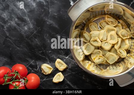 Freshly cooked tortellini are in a metal colander. The colander stands on a dark marble plate in the kitchen. There are still tomatoes on the plate. T Stock Photo