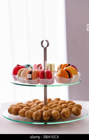 A beautiful two-tiered glass stand showcases colorful macarons nestled in paper cups, accompanied by a circle of walnuts against a clean white backdro Stock Photo