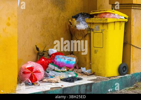 Yellow trash can with a black sign on it. The trash can is full of garbage and is located next to a wall Stock Photo