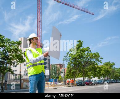 Professional young male construction worker with safety helmet and reflective vest checking work plan at a construction site with building progress in Stock Photo
