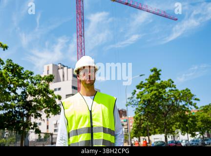 Professional young male construction worker with safety helmet and reflective vest at a construction site with building progress in the background Stock Photo