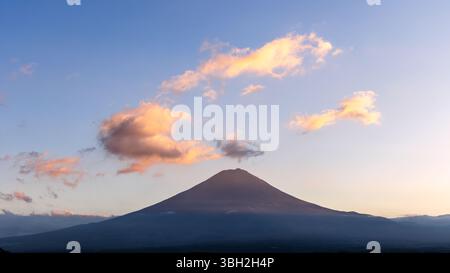 Mount Fuji stands in perfect symmetry beneath a vast sky, with sunset-lit clouds drifting above, casting a warm glow over the volcanic slopes in the e Stock Photo