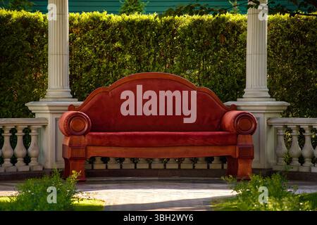 Luxurious red vintage-style sofa with wooden details placed in a classical garden pavilion. Surrounded by white columns, balustrades, and greenery. Stock Photo