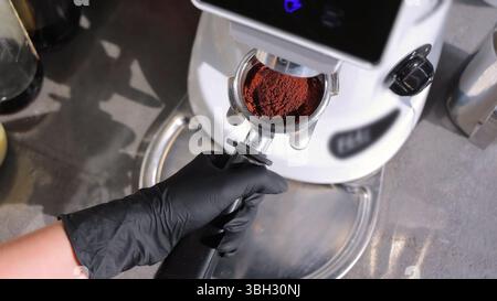 Barista Grinding Fresh Coffee Beans for Espresso. A barista wearing a black glove holds a portafilter with freshly ground coffee beneath a grinder in Stock Photo
