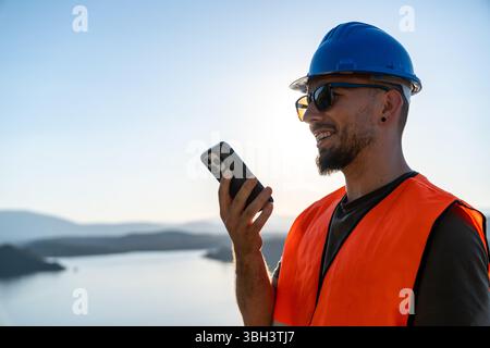 Engineer wearing hardhat and safety vest using smartphone at construction site Stock Photo