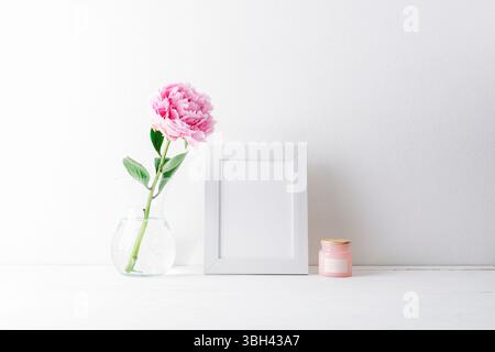 Minimal still life with pink peony in glass vase, blank picture frame and candle on white table against bright wall. Mockup. Stock Photo