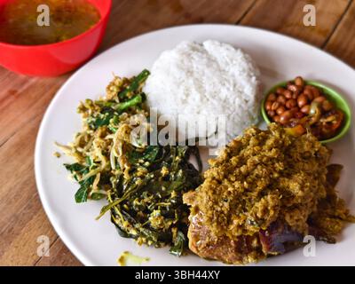 A roasted Betutu chicken rice dish featuring spiced chicken, sautéed greens, bean sprouts, fried peanuts, sambal, and spiced herb soup Stock Photo