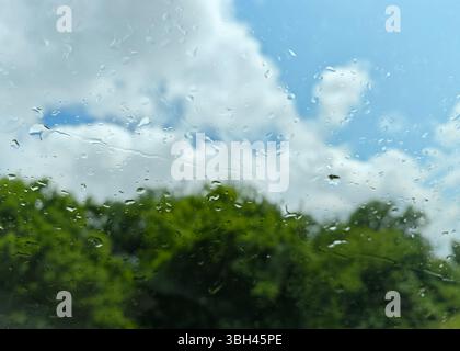 Raindrops create a pattern on a car window, framing a clear blue sky with fluffy clouds above vibrant green foliage. The scene captures a blend of rai Stock Photo