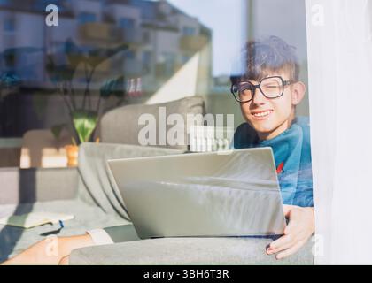 Happy student in glasses sitting with laptop and looking blinded by the sun through the window, student studying online. High quality photo Stock Photo