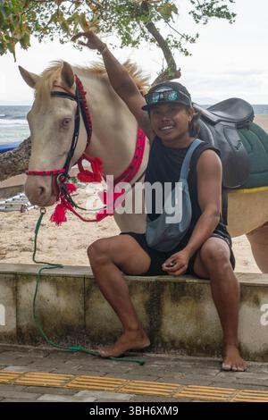 Gili Trawangan, Indonesia - Jan 4, 2025: Man are sitting on a wall next to a white horse. He is wearing a black shirt and shorts Stock Photo