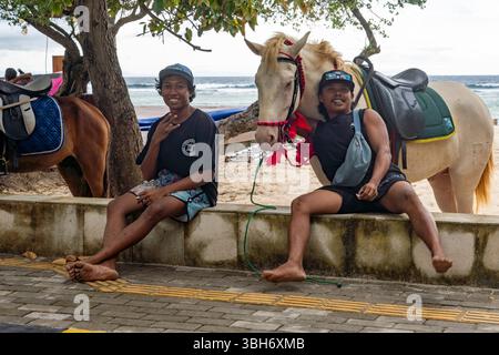 Gili Trawangan, Indonesia - Jan 4, 2025: Two young people are sitting on a wall next to a white horse. One of them is wearing a black shirt and shorts Stock Photo