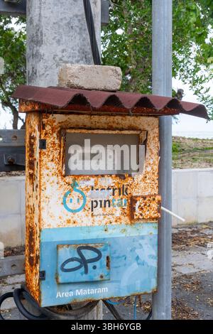 Gili Trawangan, Indonesia - Jan 4, 2025: Rusty old electricity meter with the word "Listrik Pintar" on it. The meter is located on a pole next to a tr Stock Photo