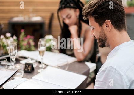 A young diverse couple on a romantic date, sitting at a table on an outdoor restaurant terrace and reading the menu to choose their meal. Fine dining Stock Photo