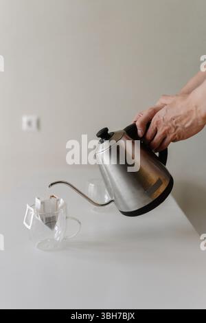 Vertical cropped shot of unrecognizable man preparing drip coffee bag by pouring hot water from stainless steel kettle into clear glass cup placing on Stock Photo