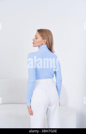 Blonde woman stands with her back to the camera, wearing a light blue long-sleeve turtleneck sweater and white high-waisted jeans. She has a relaxed Stock Photo