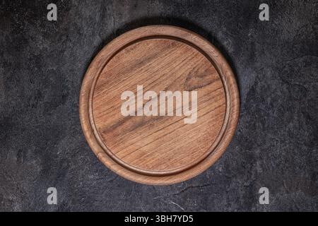 Close-up of round wooden cutting board with visible knife marks, centered on dark slate surface in studio lighting Stock Photo