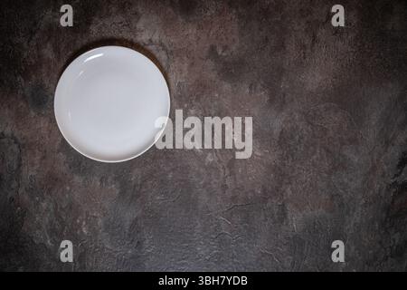 Empty white porcelain plate positioned top left on dark slate surface, ample space for text or design Stock Photo