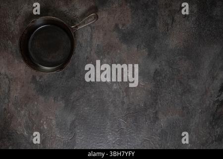 Small empty single-piece cast iron frying pan positioned top left on dark slate surface, ample space for text or design Stock Photo