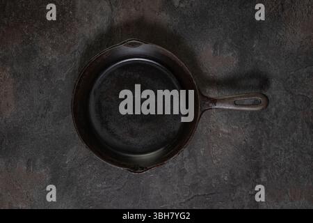 Small empty single-piece cast iron frying pan without handle, centered on dark slate surface in studio lighting. For food styling photography. Stock Photo
