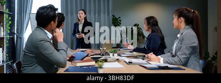 Adult Asian businesswoman leads a presentation to a diverse team during a corporate meeting in a modern office setting. Team members collaborate brain Stock Photo