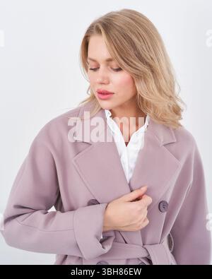 Blonde woman wearing a light mauve trench coat over a white blouse, with her eyes gently closed, exuding a serene demeanor and softly styled loose Stock Photo