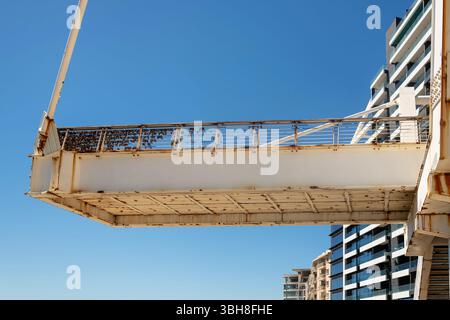 Bottom view of a rusty pedestrian bridge with love padlocks on the railing, modern buildings in the background, and a clear blue sky. Stock Photo