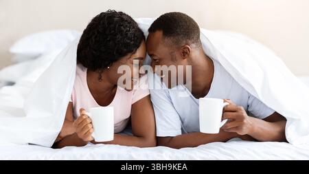 Romantic black couple laying under blanket, drinking tea Stock Photo