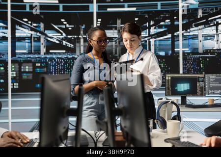 Team of technicians using tablet in server farm, analyzing data. Women working in data center examining infrastructure using device, ensuring system integrity and security Stock Photo