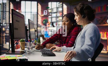 Call center team leader going over paperwork files next to colleague at PC desk. Customer support manager explaining company documentation to intern accommodating with job, camera B Stock Photo