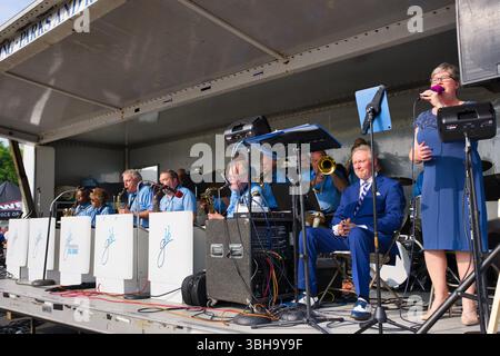 A big band, predominantly in blue uniforms, performs on a stage. A woman in a blue dress sings into a microphone.  The scene is brightly lit, outdoors Stock Photo
