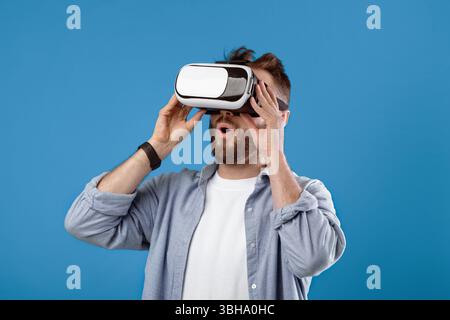 Cool millennial guy experiencing virtual reality in VR headset on blue studio background Stock Photo