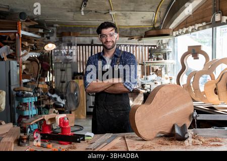 Smiling luthier standing with arms crossed in workshop, surrounded by guitar parts and tools. Concept of craftsmanship, woodworking, handmade instrume Stock Photo